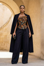 Woman in a stylish outfit with a long black coat, patterned top, and wide-leg pants standing in front of a stone archway.